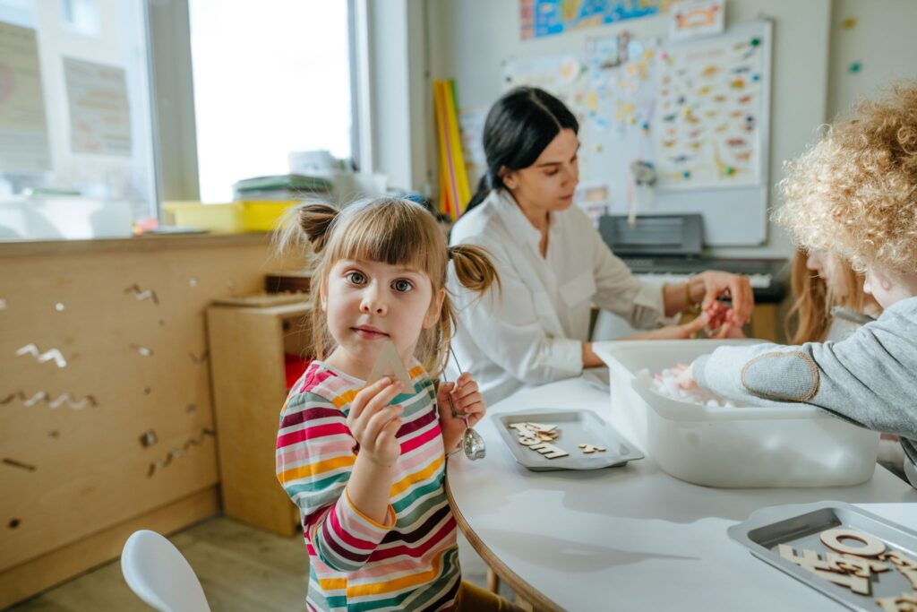 Children playing in kindergarten with teacher. Portrait of adorable 4 years old girl with wooden letter A looking at camera. Education concept. Selective focus.